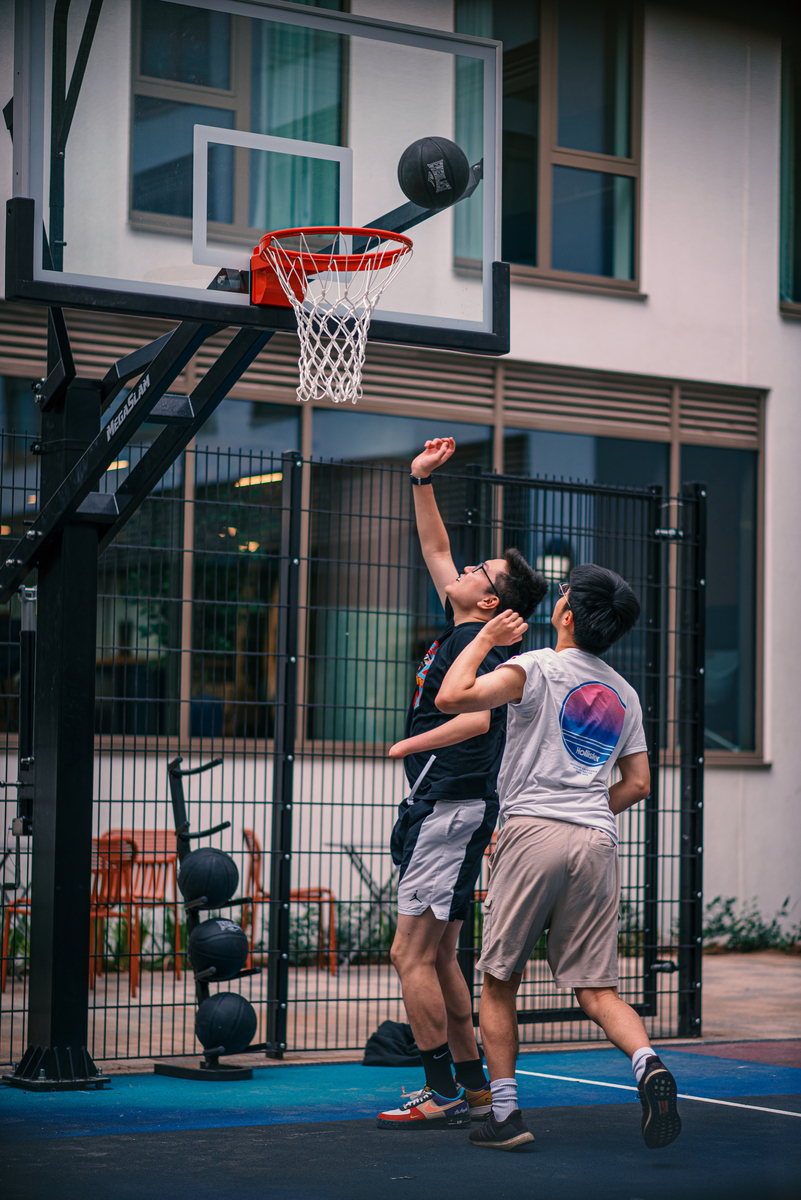 Two people playing basketball outdoors. One is attempting a layup while the other defends. A building and fence are in the background. The action is dynamic, with the ball near the hoop. Both players are wearing shorts and athletic shirts.